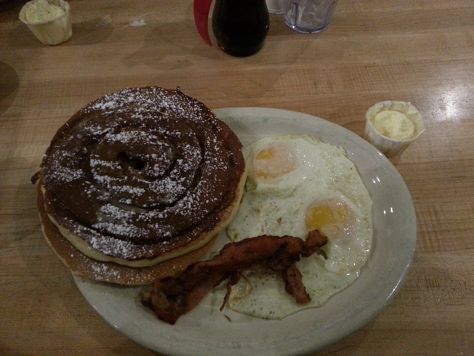 A stacked breakfast of pancakes, eggs, and bacon on a diner plate.